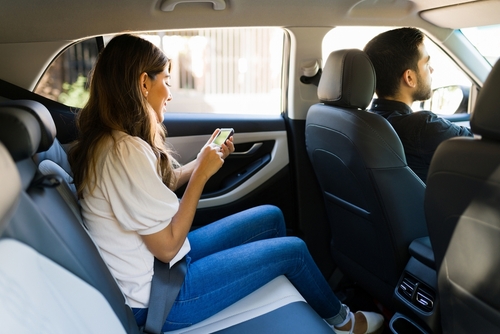 Woman sitting in the back seat of a rideshare car looking at her phone while the driver focuses on the road.