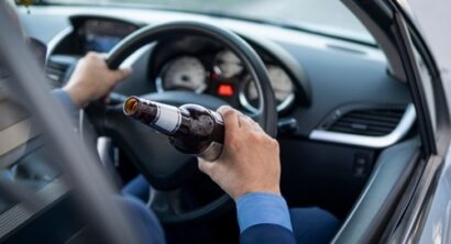 Driver holding a beer bottle while operating a car, showing the steering wheel and dashboard