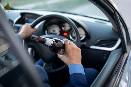 Driver holding a beer bottle while operating a car, showing the steering wheel and dashboard