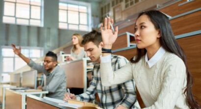 Female international student in a lecture hall raising her hand to ask a question while classmates listen and take notes.