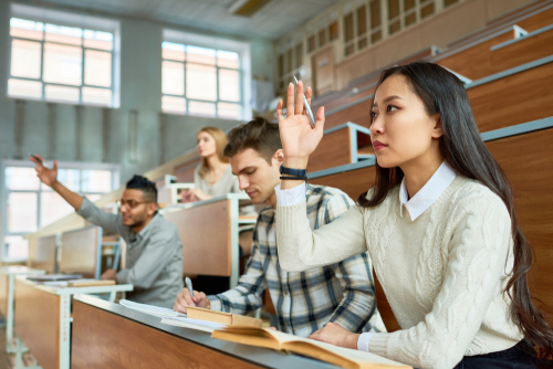 Female international student in a lecture hall raising her hand to ask a question while classmates listen and take notes.