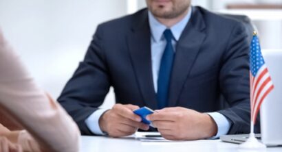 Immigration lawyer seated at a desk with a small American flag, holding documents while speaking with a client about legal status.