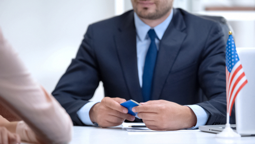 Immigration lawyer seated at a desk with a small American flag, holding documents while speaking with a client about legal status.