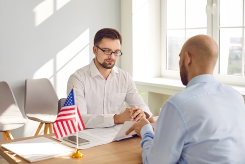 Man meeting with immigration officer across a desk, small American flag on table