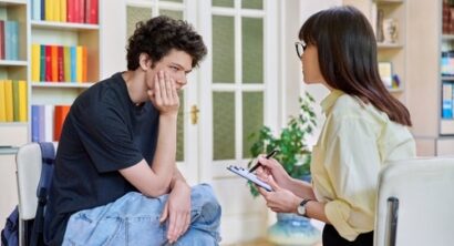 Teenage boy seated with a thoughtful expression during a counseling session as a female therapist takes notes in a brightly lit office.