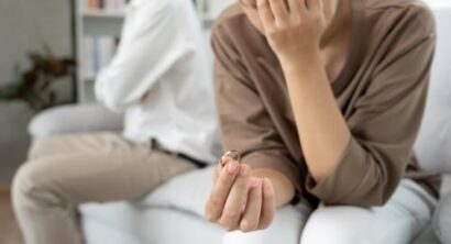 Woman holding a wedding ring while sitting on a couch with her spouse in the background after a difficult conversation about divorce.